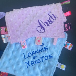 May include: Two square baby security blankets with a dimpled texture. The top blanket is light pink with the name Andi embroidered in dark purple. The bottom blanket is light blue with the name Ioannis Xristos embroidered in dark blue. Both feature colourful patterned ribbon tags along the edges.