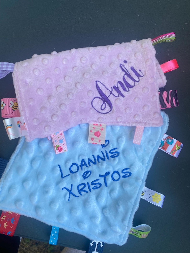 May include: Two square baby security blankets with a dimpled texture. The top blanket is light pink with the name Andi embroidered in dark purple. The bottom blanket is light blue with the name Ioannis Xristos embroidered in dark blue. Both feature colourful patterned ribbon tags along the edges.