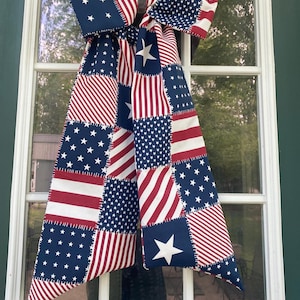 Puede incluir: Un lazo decorativo patriótico con un diseño de patchwork de la bandera estadounidense. El lazo está hecho de tela con patrones alternos de rayas rojas y blancas, campos azules con estrellas blancas y una gran estrella blanca. Cuelga de un marco de ventana blanco.