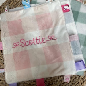 May include: A square plush security blanket featuring a soft white and light pink buffalo check pattern. The name Scottie is embroidered in pink cursive script across the centre. The blanket is bordered by various colourful ribbon tags.