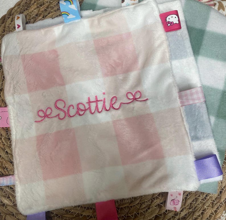 May include: A square plush security blanket featuring a soft white and light pink buffalo check pattern. The name Scottie is embroidered in pink cursive script across the centre. The blanket is bordered by various colourful ribbon tags.