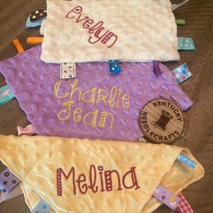 May include: Three rectangular and triangular sensory blankets with dimpled textures in cream, lavender, and pale yellow. Each blanket features embroidered names: Evelyn, Charlie Jean, and Melina, along with various colourful patterned ribbon tags.