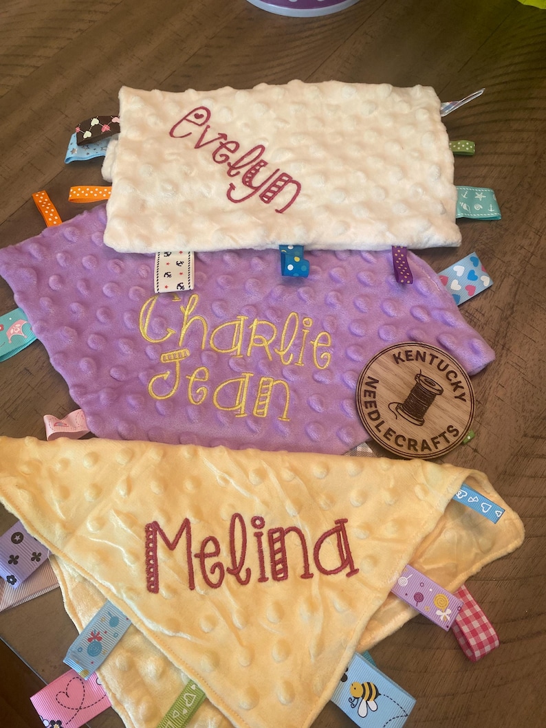 May include: Three rectangular and triangular sensory blankets with dimpled textures in cream, lavender, and pale yellow. Each blanket features embroidered names: Evelyn, Charlie Jean, and Melina, along with various colourful patterned ribbon tags.