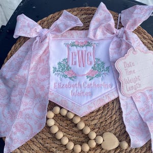 May include: A personalized baby banner with pink floral accents and the initials "EWC" embroidered in pink. The banner includes the name "Elizabeth Catherine Waites" and a tag for recording birth details. The banner is adorned with pink floral patterned ribbons.