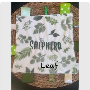 May include: A square baby security blanket featuring a white fabric base with a green leaf pattern. The name Shepherd is embroidered in dark green letters across the centre. Multiple colourful ribbon tags in various shades of green and white are attached to the edges of the blanket.