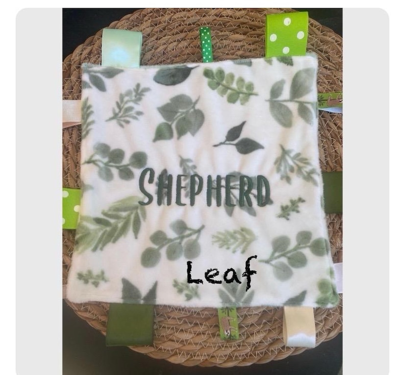 May include: A square baby security blanket featuring a white fabric base with a green leaf pattern. The name Shepherd is embroidered in dark green letters across the centre. Multiple colourful ribbon tags in various shades of green and white are attached to the edges of the blanket.
