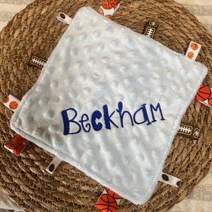 May include: A square light blue minky fabric security blanket with a dimpled texture. The name Beckham is embroidered in dark blue thread across the centre. The edges feature multiple fabric ribbon tags with basketball and football patterns.
