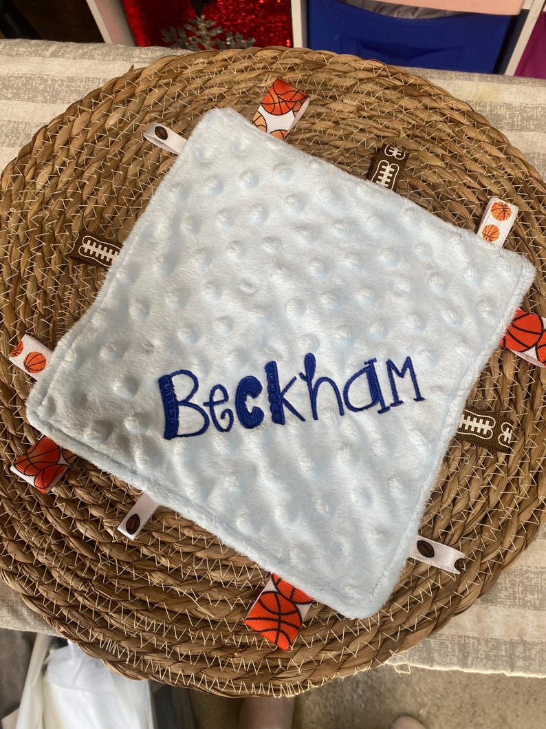 May include: A square light blue minky fabric security blanket with a dimpled texture. The name Beckham is embroidered in dark blue thread across the centre. The edges feature multiple fabric ribbon tags with basketball and football patterns.