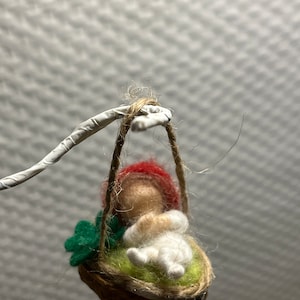 Könnte beinhalten: Eine kleine Filzpuppe mit rotem Hut und weißer Kleidung sitzt in einer Nussschale. Die Wiege hängt an einer Schnur und hat grüne Filzblätter am Rand.