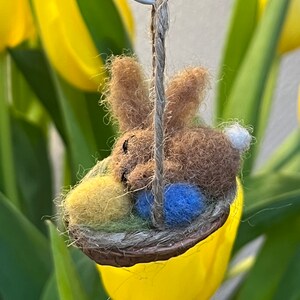 Könnte beinhalten: Ein braunes Filz-Hasen-Ornament mit weißem Schwanz sitzt in einem kleinen gewebten Korb mit einem gelben und blauen Ei. Das Ornament hängt an einer Juteschnur und einem weißen Draht. Der Hintergrund zeigt gelbe Tulpen.