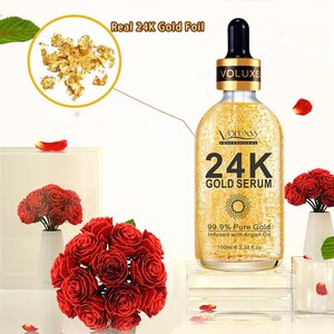 Op de afbeelding: Een gouden fles met een druppelaar en een gouden etiket met de tekst "VOLUXSS PROFESSIONAL 24K GOLD SERUM 99.9% Pure Gold Infused with Argan Oil 100ml e 3.38 fl.oz". De fles staat op een wit oppervlak met rode rozen op de voorgrond.