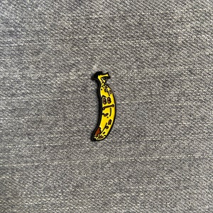以下が含まれることがあります： 茶色の斑点のあるバナナの黄色いエナメルピン。バナナは黒い輪郭で漫画風に描かれています。