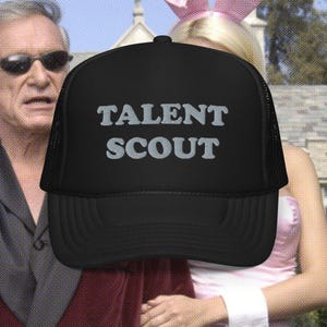 Peut inclure: Casquette de camionneur noire avec un filet blanc à l'arrière. La casquette a le texte "TALENT SCOUT" brodé à l'avant en fil argenté.