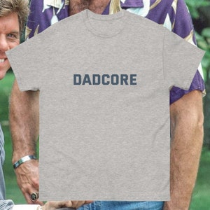 May include: A grey t-shirt featuring the word "DADCORE" in dark blue block lettering. The shirt has a crew neck and short sleeves. The t-shirt is a casual style.