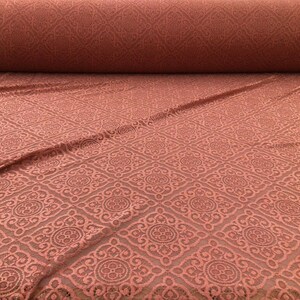 May include: A roll of coral-colored lace fabric with an intricate geometric pattern. The fabric is displayed flat, with a portion rolled up in the background, showcasing the detailed design and texture.
