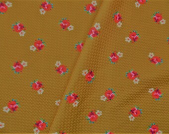 May include: Mustard-colored fabric with a repeating pattern of small red and white floral clusters. The fabric has a textured, woven appearance, and a fold is visible, adding depth to the image. The floral design is evenly spaced across the surface.