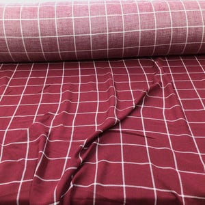 May include: Red and white plaid fabric with a large white grid pattern. The fabric is folded and draped, showing the texture and color of the material.