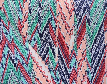 May include: Close-up of a fabric with a colorful chevron pattern. The design features a repeating zigzag pattern in shades of navy blue, coral, light green, and peach. The fabric appears to be folded, creating a layered effect.