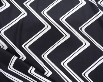 May include: Close-up of a black fabric with a repeating white geometric pattern. The pattern consists of white outlined, angular shapes arranged in a zig-zag design. The fabric has a slight sheen and is folded in places.