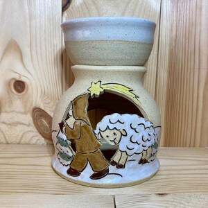 May include: A ceramic candle warmer with a beige glaze and a scene of a person carrying a tree and a sheep. The scene is painted in brown, green, yellow, and white.