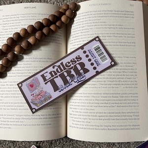 Könnte beinhalten: Ein violettes und braunes Lesezeichen mit dem Text "Endless TBR Book Club" und einem Ticket mit der Aufschrift "Admit One". Das Lesezeichen zeigt einen Stapel Bücher mit einer Skeletthand, die ein Herz hält.