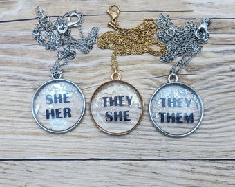 May include: Three silver necklaces with round glass pendants. The pendants have silver, gold, and silver frames. The pendants have glitter inside and black text that reads "She Her", "They She", and "They Them".