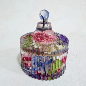 Peut inclure: Un bocal décoratif transparent avec un couvercle, rempli de fleurs séchées colorées. Le bocal a un design côtelé et un bouton arrondi transparent sur le dessus. Les fleurs comprennent des nuances de rose, de bleu, de violet et de vert.
