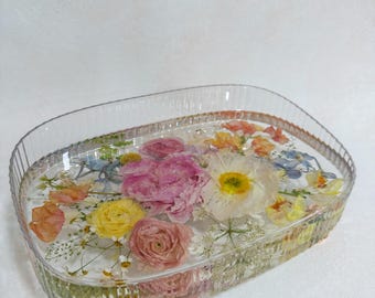 May include: Clear, oval-shaped resin tray with embedded dried flowers in shades of pink, yellow, orange, and white. The tray has a decorative, ridged edge and rests on small, gold-colored feet. A unique decorative piece.