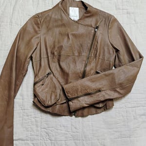 Peut inclure: Veste en cuir marron avec fermeture &eacute;clair asym&eacute;trique et deux poches zipp&eacute;es. La veste a un design sans col et des manches longues. Le cuir a un aspect l&eacute;g&egrave;rement vieilli.
