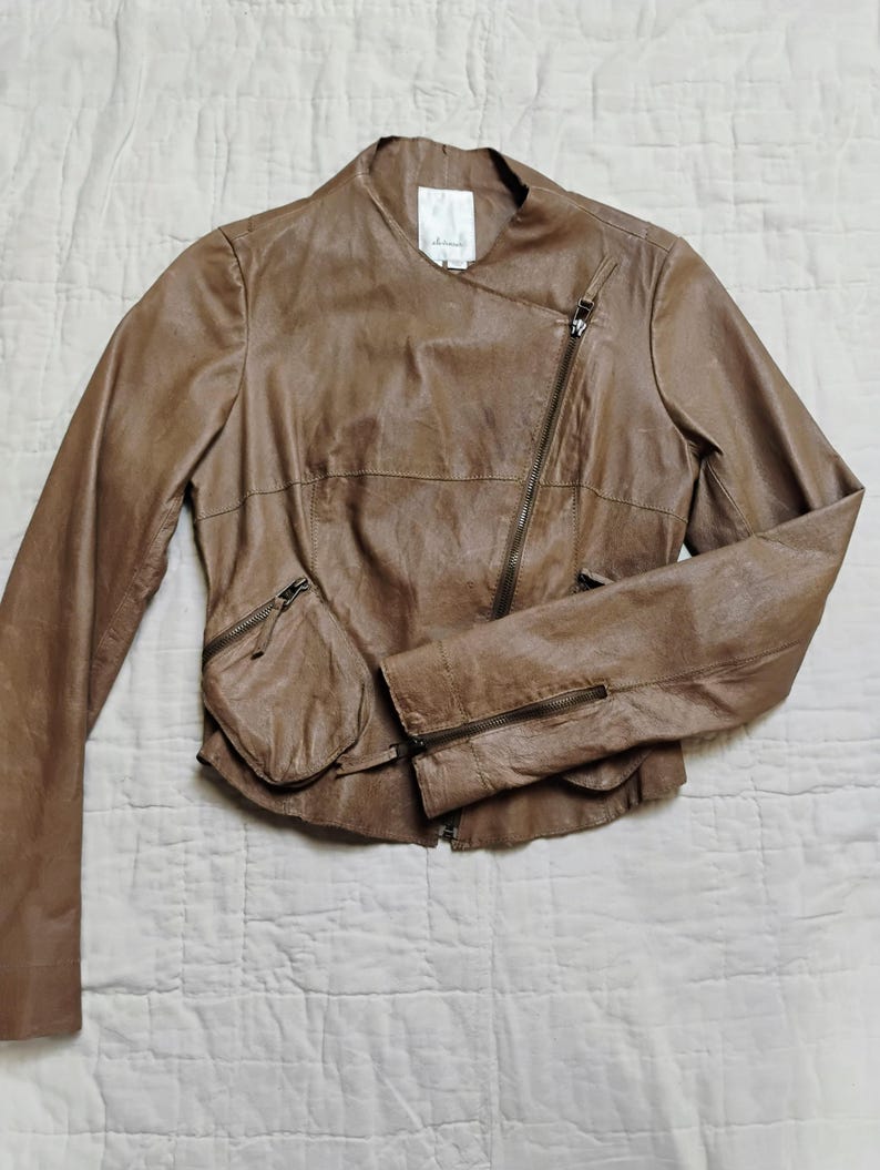 Peut inclure: Veste en cuir marron avec fermeture &eacute;clair asym&eacute;trique et deux poches zipp&eacute;es. La veste a un design sans col et des manches longues. Le cuir a un aspect l&eacute;g&egrave;rement vieilli.