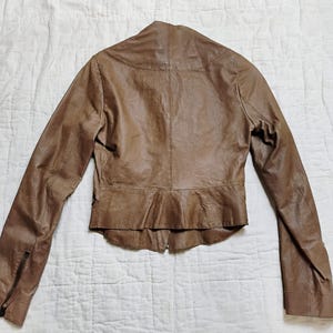 Peut inclure: Veste en cuir marron avec des manches longues et une fermeture &eacute;clair sur chaque manche. La veste a une silhouette ajust&eacute;e avec un ourlet &agrave; volants et une fermeture &eacute;clair &agrave; l'avant. La veste est en cuir souple et textur&eacute;.