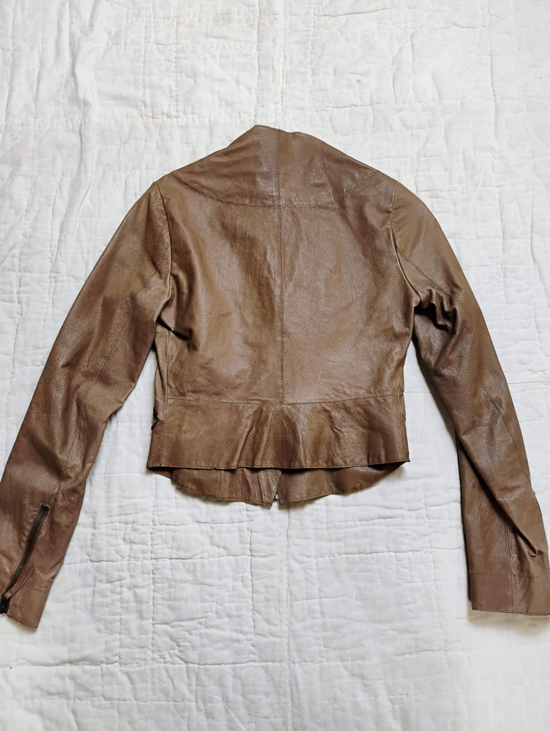 Peut inclure: Veste en cuir marron avec des manches longues et une fermeture &eacute;clair sur chaque manche. La veste a une silhouette ajust&eacute;e avec un ourlet &agrave; volants et une fermeture &eacute;clair &agrave; l'avant. La veste est en cuir souple et textur&eacute;.