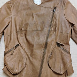 Peut inclure: Veste en cuir marron clair avec fermeture &eacute;clair asym&eacute;trique et deux poches avant. Le col est absent et la coupe est ajust&eacute;e. Le cuir semble souple et vieilli, lui donnant un aspect vintage et &eacute;l&eacute;gant.