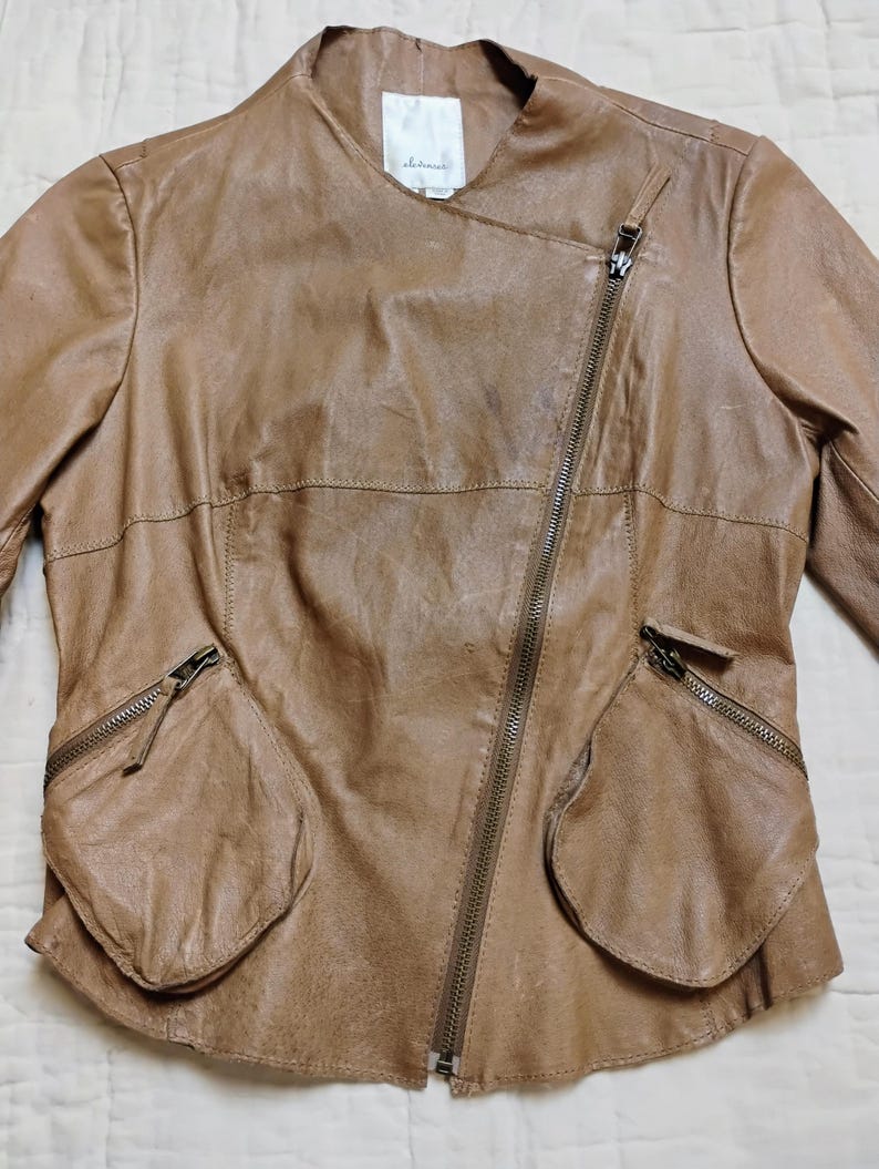Peut inclure: Veste en cuir marron clair avec fermeture &eacute;clair asym&eacute;trique et deux poches avant. Le col est absent et la coupe est ajust&eacute;e. Le cuir semble souple et vieilli, lui donnant un aspect vintage et &eacute;l&eacute;gant.