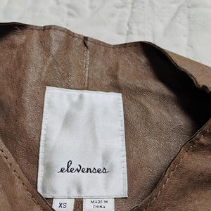 Peut inclure: Gros plan sur un v&ecirc;tement en cuir marron avec une &eacute;tiquette blanche. L'&eacute;tiquette indique "elevenses" en cursive, avec "XS" et "MADE IN CHINA" imprim&eacute; en dessous. Le cuir a un aspect textur&eacute;.