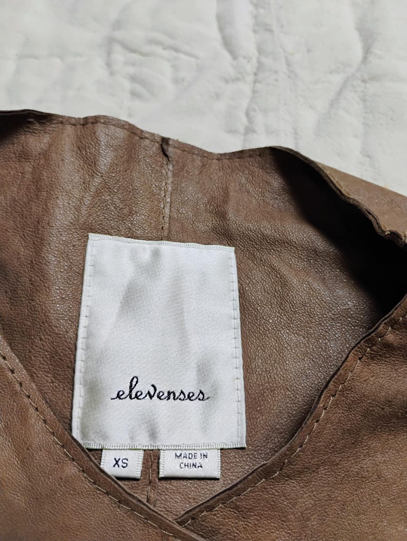 Peut inclure: Gros plan sur un v&ecirc;tement en cuir marron avec une &eacute;tiquette blanche. L'&eacute;tiquette indique "elevenses" en cursive, avec "XS" et "MADE IN CHINA" imprim&eacute; en dessous. Le cuir a un aspect textur&eacute;.