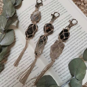 Puede incluir: Tres llaveros con dijes de piedra natural envueltos en cuerda marrón. Cada dije está atado con una borla marrón. Los dijes son un cristal transparente, una piedra gris oscura y una piedra rosa claro.