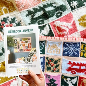 Puede incluir: Una mano sostiene un folleto titulado "THE HEIRLOOM ADVENT CALENDAR" con una colorida manta de punto en el fondo. La manta presenta cuadrados con varios diseños en tonos de rosa, verde, blanco y azul. El folleto describe un calendario de adviento tejido.