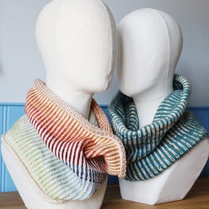 May include: Two knitted cowls displayed on mannequin heads. One cowl has a gradient of colours, including red, orange, yellow, green, and blue. The other cowl is teal and white striped. Both are ribbed knit.