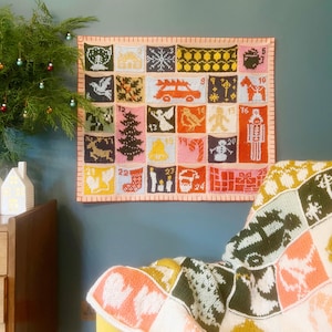 Pode incluir: Um calendário do advento de malha colorido com 24 bolsos numerados, cada um com um design temático de Natal único. O calendário é emoldurado com uma borda laranja clara e pendurado em uma parede azul-petróleo. Uma manta de malha combinando está sobre uma cadeira amarela.