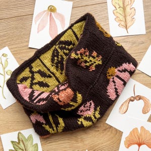 May include: A dark brown knitted cowl with floral and leaf patterns in pink, yellow, and orange. The cowl is surrounded by watercolour paintings of flowers and leaves on a wooden surface.