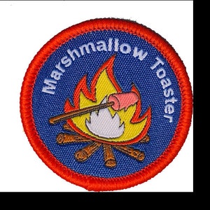 Może przedstawiać: Naszywka haftowana w kolorze niebieskim i czerwonym z napisem "Marshmallow Toaster" i kreskówkowym obrazkiem pianki na patyku nad ogniskiem.