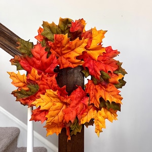 May include: A fall-themed wreath composed of vibrant, multi-colored maple leaves in shades of red, orange, and yellow. The wreath is arranged in a circular shape, perfect for seasonal home decor. The leaves are clustered together, creating a full and textured appearance.