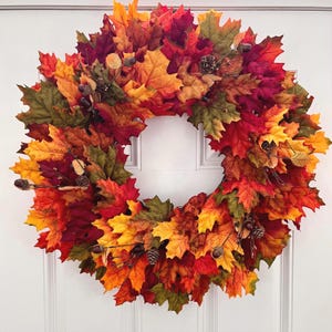May include: A circular wreath composed of artificial autumn leaves in shades of red, orange, yellow, and green. The foliage is interspersed with small brown acorns and miniature pinecones, creating a dense, layered texture throughout the design.