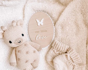 May include: A knitted beige baby toy shaped like a giraffe, a wooden oval sign with a butterfly and the words "our little love", and a pair of knitted baby booties. The items are arranged on a white textured blanket.