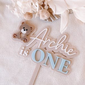 May include: A cake topper featuring a beige base with a brown teddy bear icon and the white and light blue text "Archie is ONE". The topper has a long, thin, vertical stem extending from the bottom, designed for insertion into a cake or dessert.