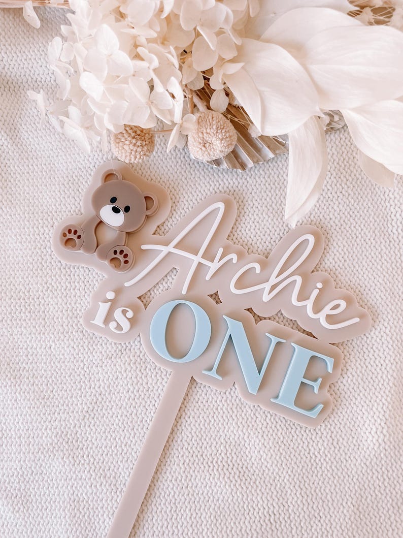 May include: A cake topper featuring a beige base with a brown teddy bear icon and the white and light blue text "Archie is ONE". The topper has a long, thin, vertical stem extending from the bottom, designed for insertion into a cake or dessert.