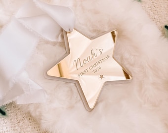 May include: A star-shaped ornament with a reflective gold mirror finish, featuring the engraved text "Noah's FIRST CHRISTMAS 2026" and a small star icon. A white silk ribbon is attached to the top for hanging, creating a festive holiday decoration.