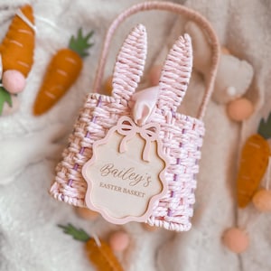 May include: A pink woven Easter basket with bunny ears and a wooden tag reading "Bailey's Easter Basket." The basket is surrounded by felt carrots and pom-poms, on a soft, white surface. A charming, festive design for Easter celebrations.