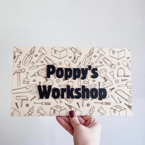 May include: A wooden sign featuring the words "Poppy's Workshop" in black, raised lettering. The sign has a light wood background with a repeating pattern of tools. A hand holds the sign. The tools include hammers, drills, and saws.